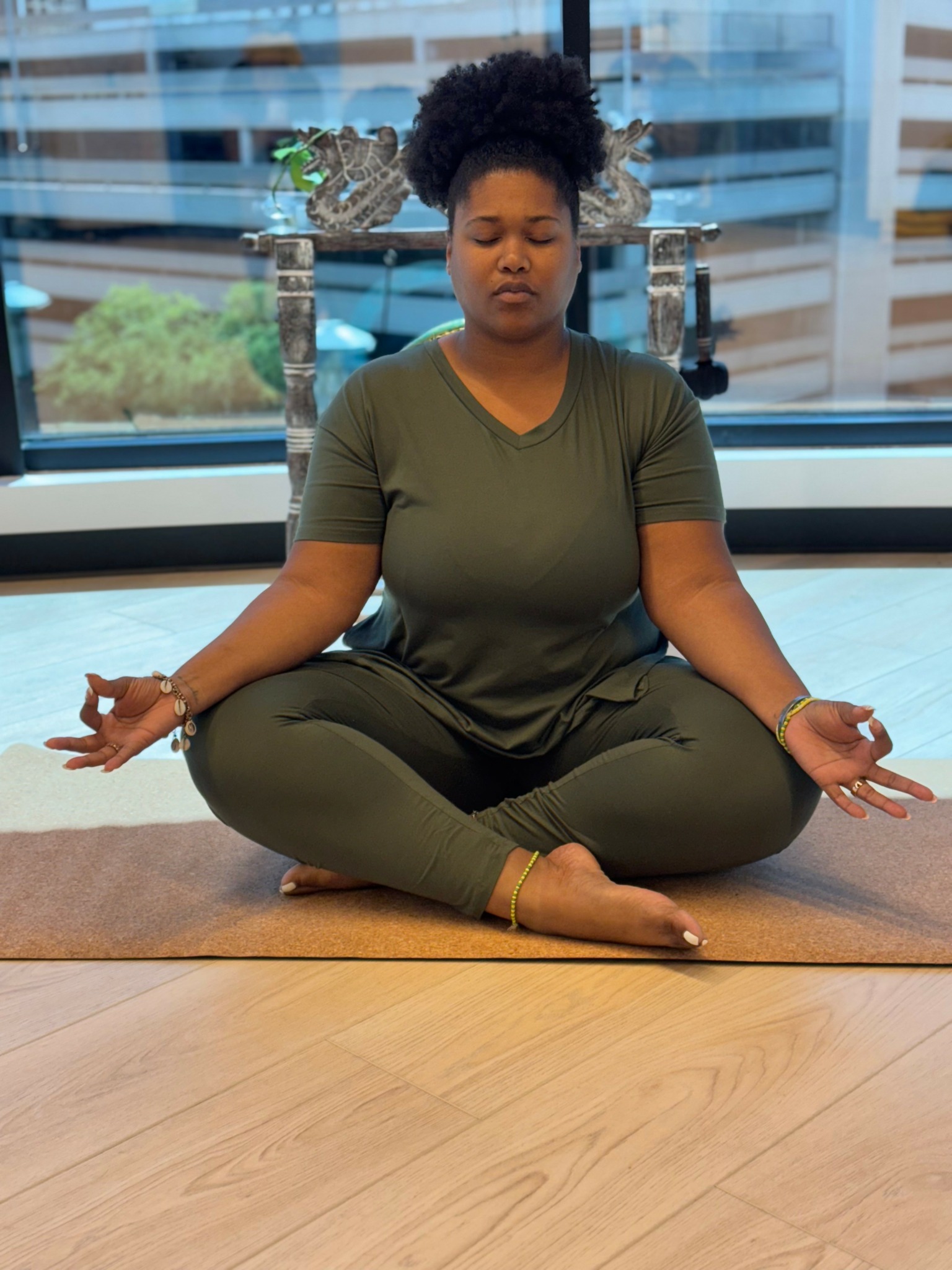 Woman sitting cross-legged on a yoga mat, meditating with eyes closed indoors, cityscape visible through large windows behind her.