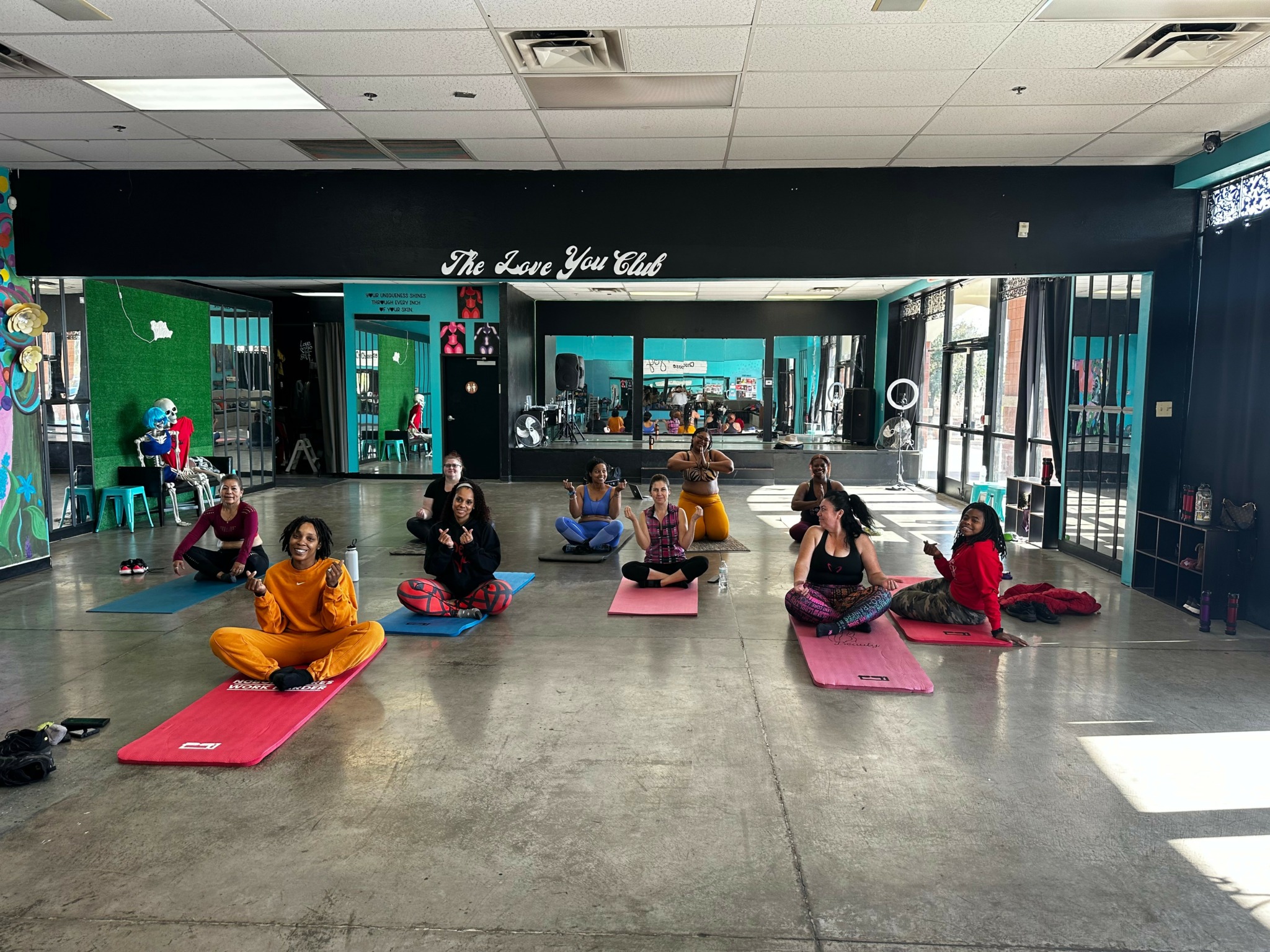 Group of people practicing yoga on mats in a studio with large windows and a sign that reads 'The Love You Club'.
