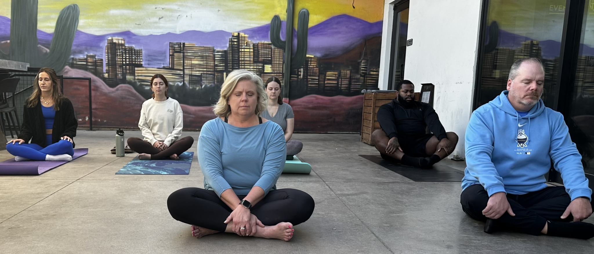 Six people sitting cross-legged on mats in a room with desert mural and windows, practicing meditation or yoga.