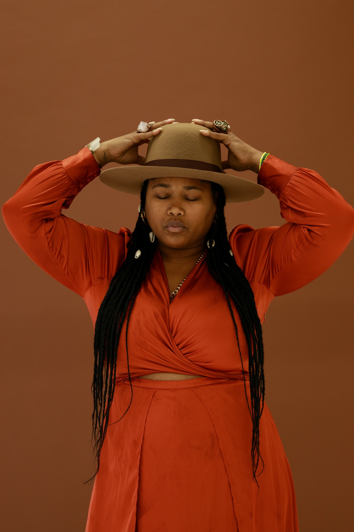 Woman with long braids wearing an orange dress and wide-brimmed hat, holding the hat with both hands, against a brown background.