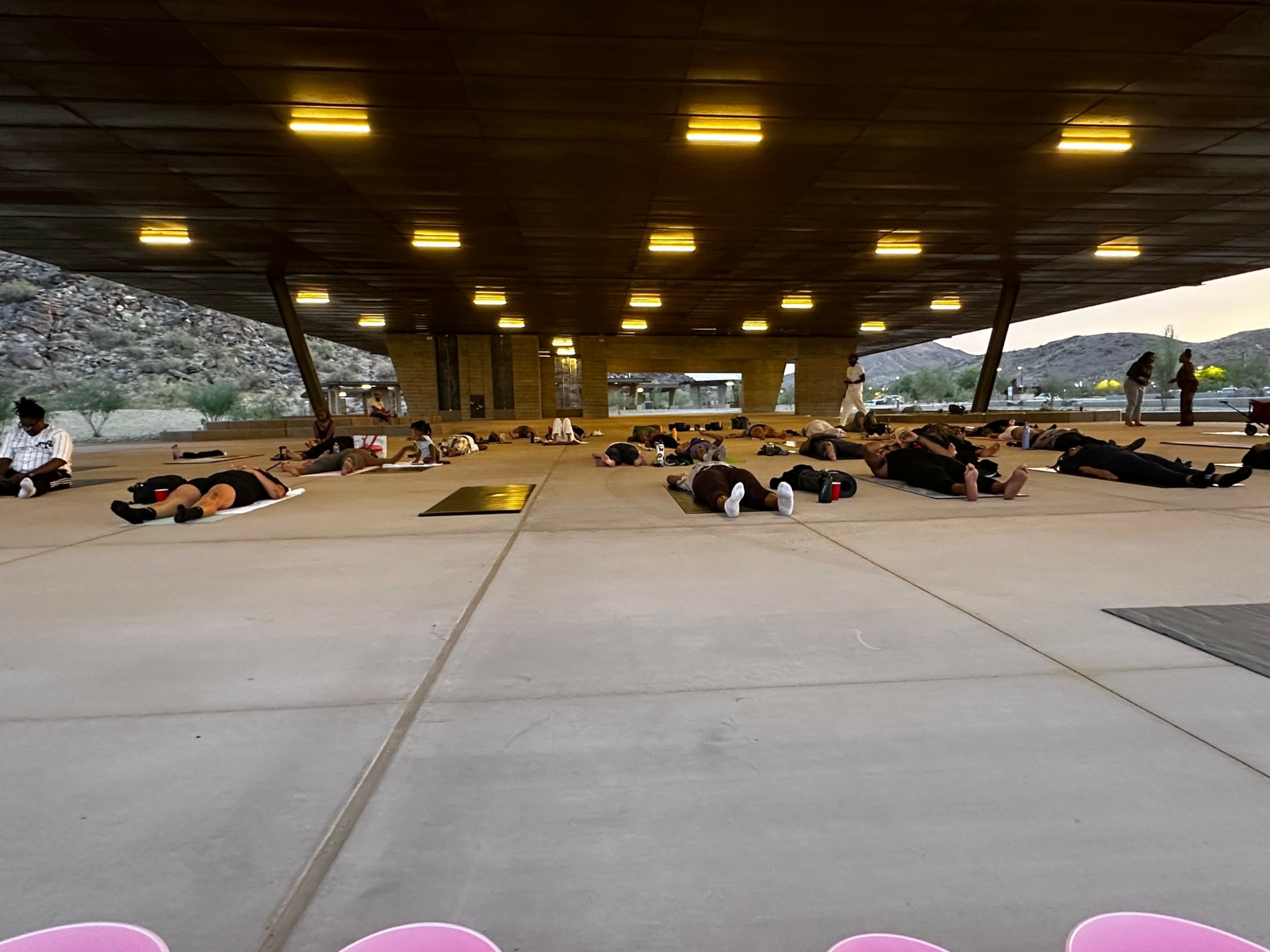 People practicing yoga or meditation outdoors under a large covered area, with a scenic landscape in the background.