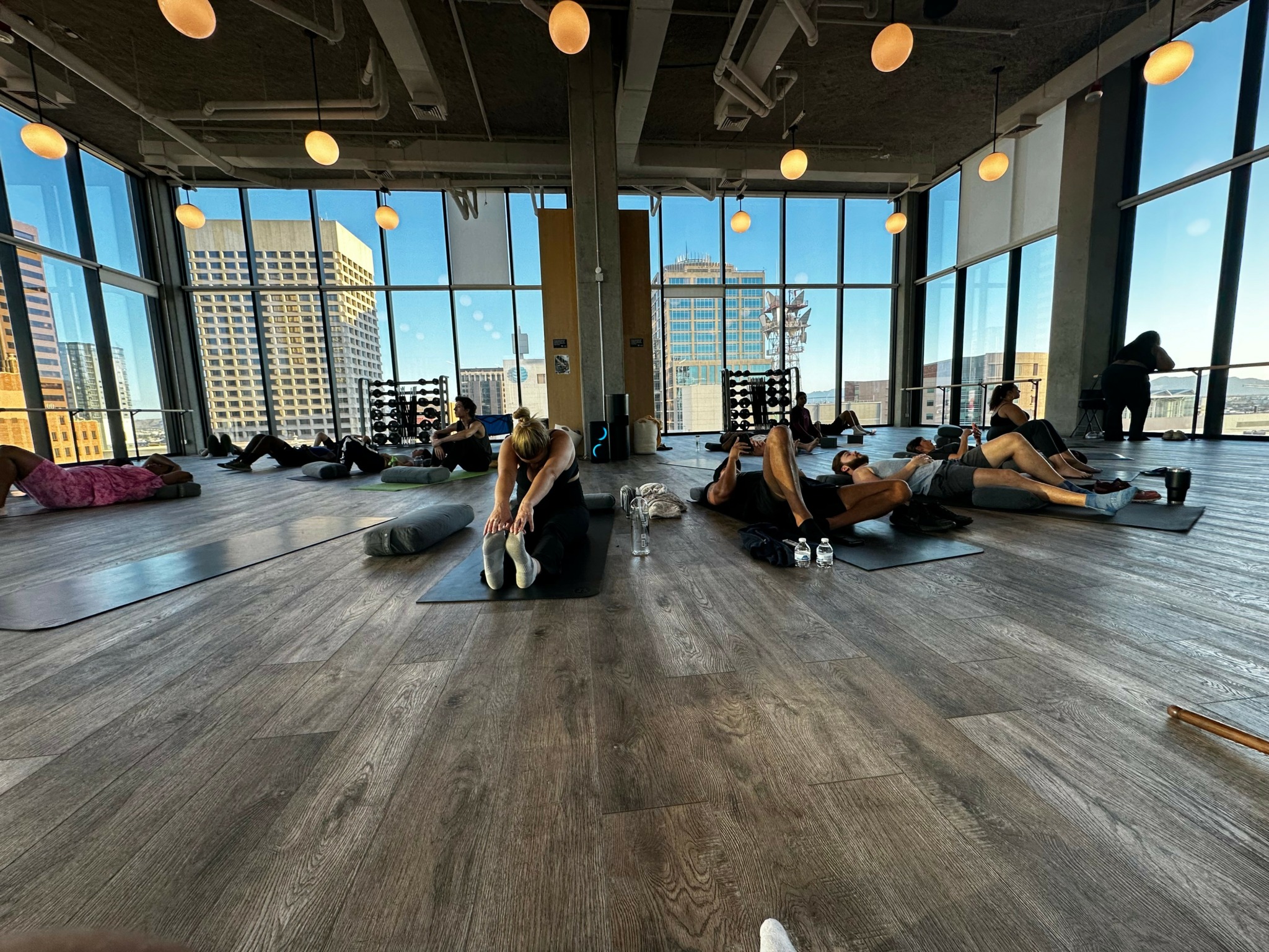People practicing yoga or stretching on mats in a bright, spacious studio with large windows and city view.