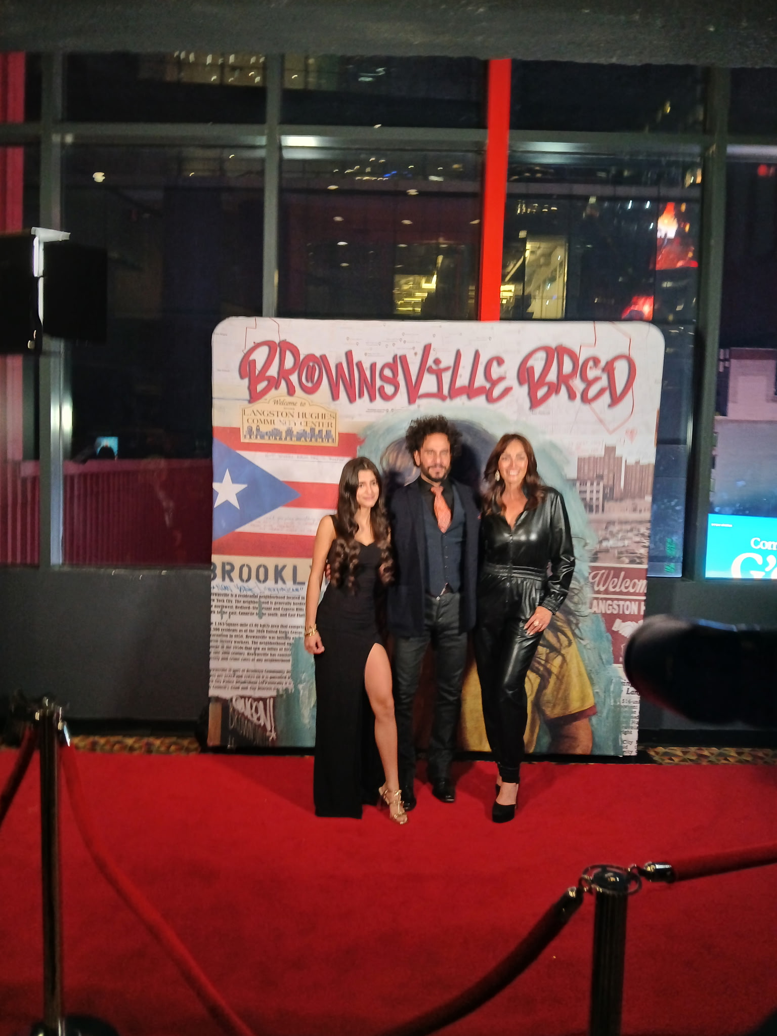 Three people standing on a red carpet in front of a promotional backdrop with text and graphics, inside a building with large windows.