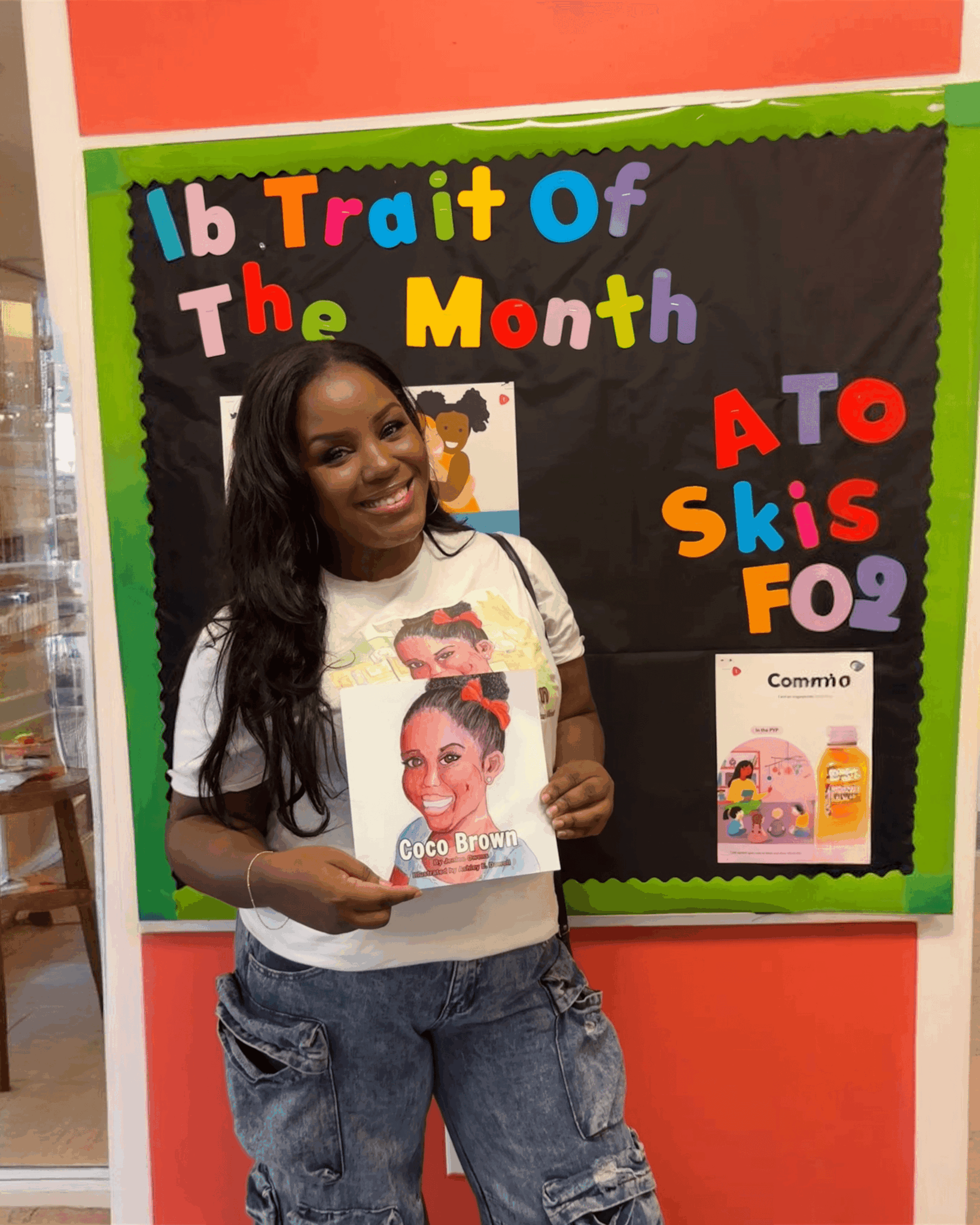 Young girl with long dark hair holding a book, standing in front of a colorful bulletin board with text and images.