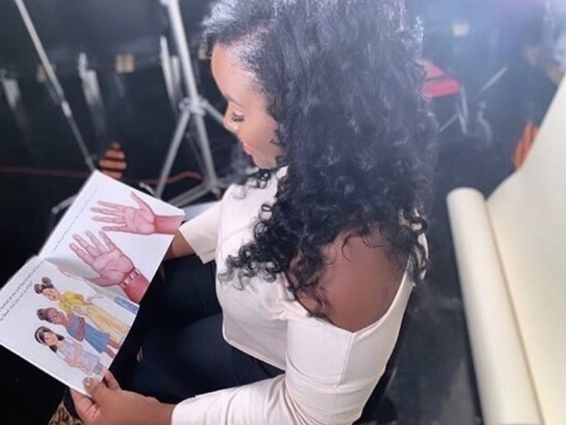 Woman with curly hair reading a colorful magazine, sitting indoors with a white chair nearby.