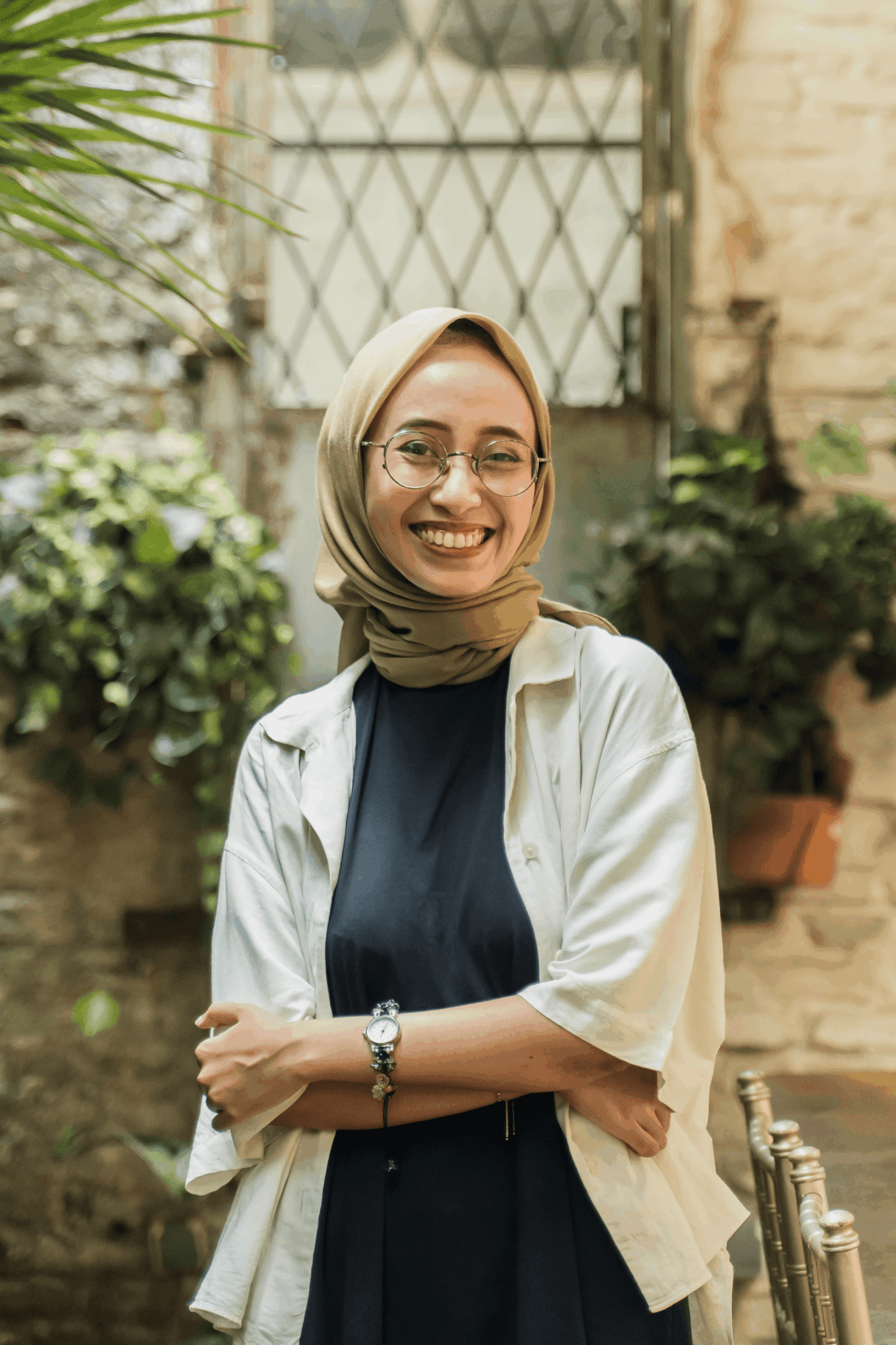 Smiling woman with glasses and headscarf standing outdoors in front of a brick wall and greenery.