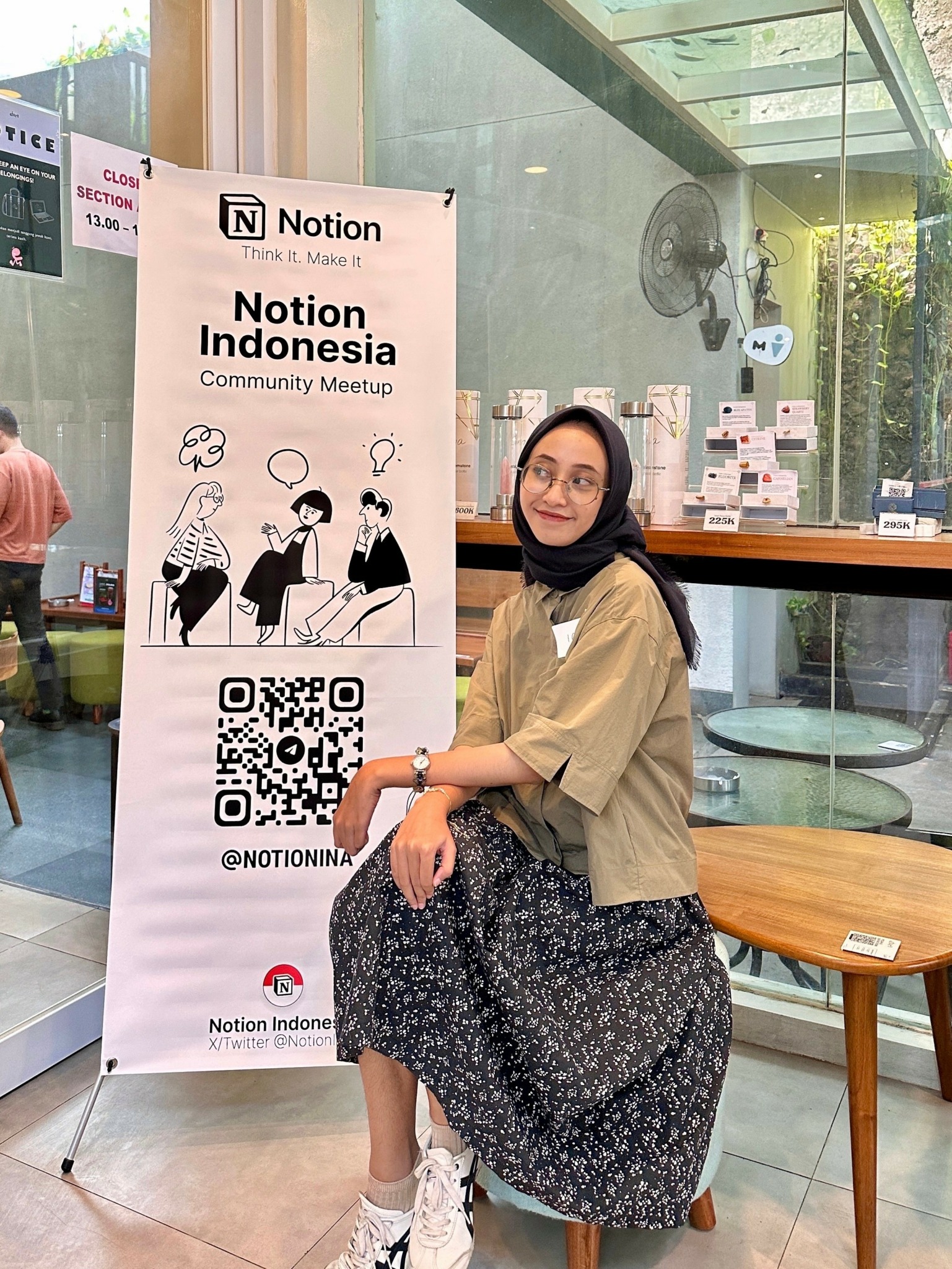 Woman sitting on a bench inside a cafe, next to a Notion Indonesia community meetup banner, with a window and plants behind.