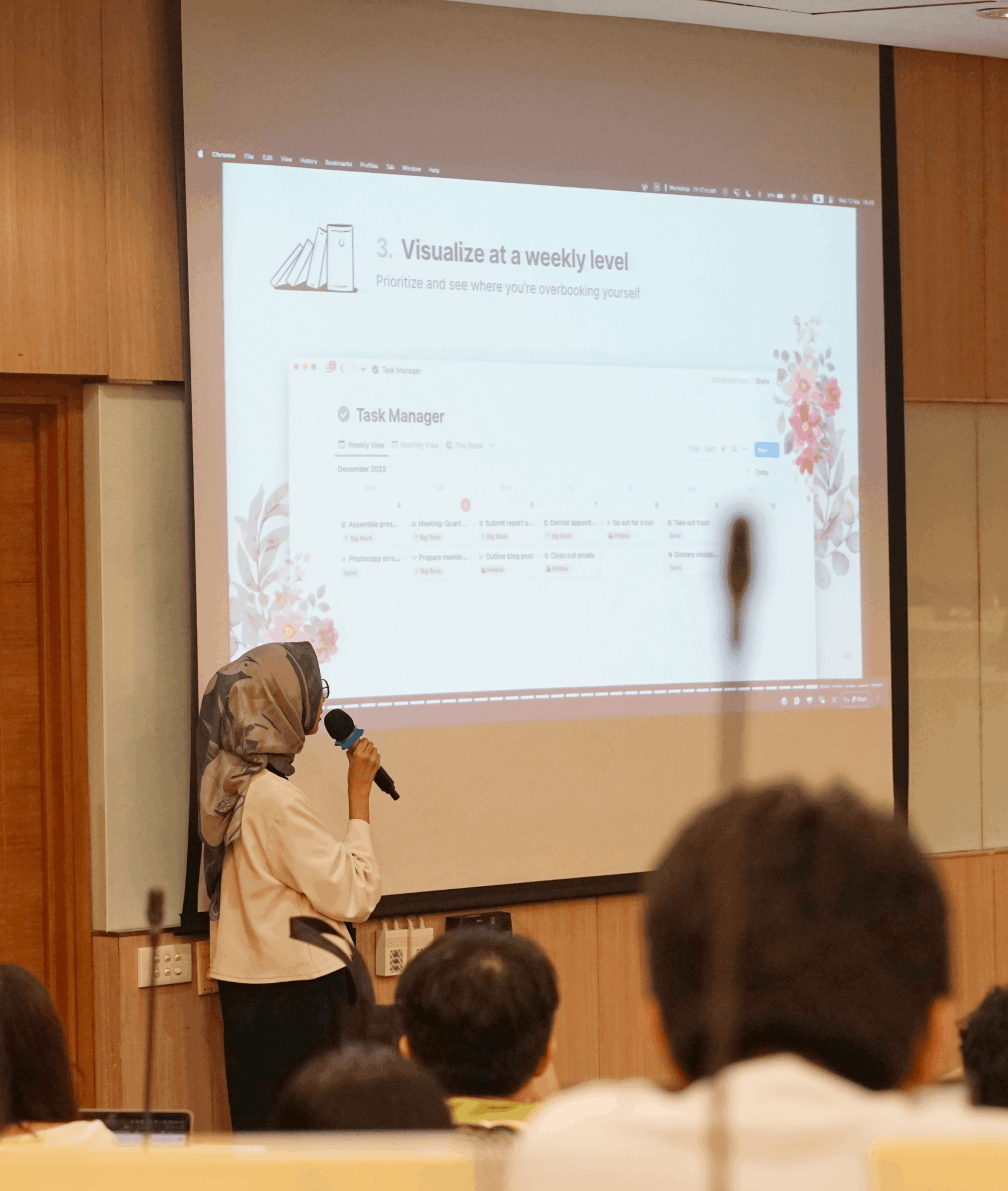 Person wearing a hijab and white shirt presenting with a microphone in front of a large screen in a conference room. Audience members are visible.