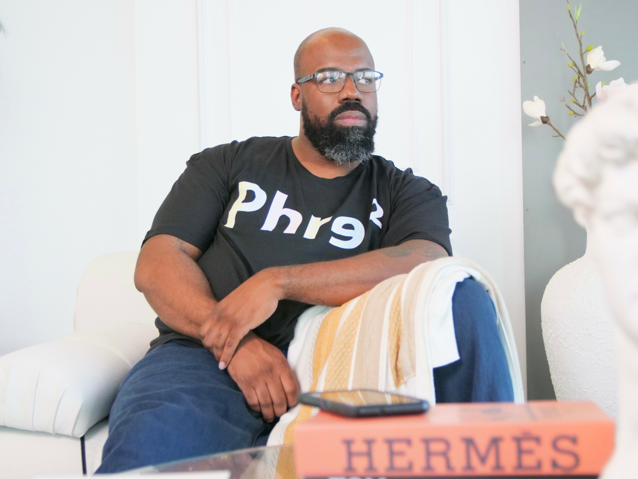Man with glasses and beard sitting on a white couch, holding a towel, with a Hermes box on table, in a bright room.