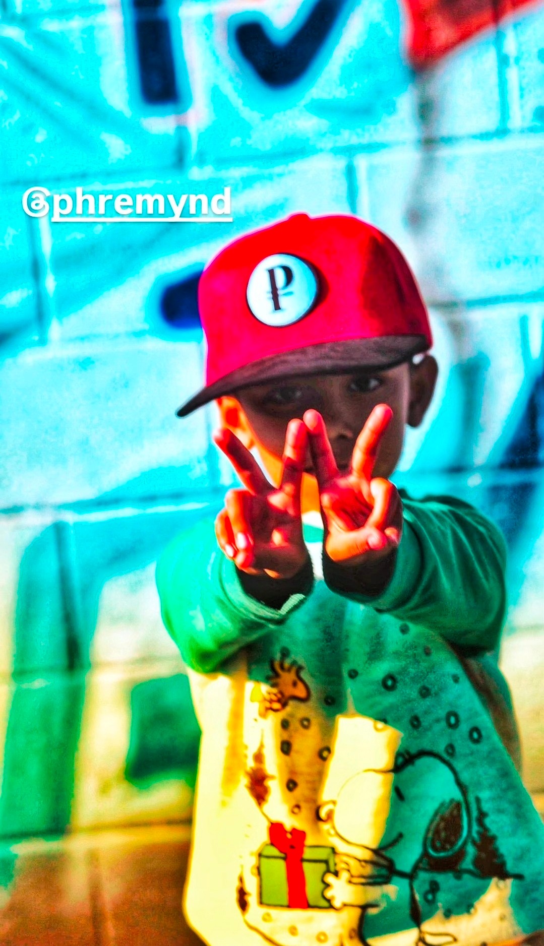 Child wearing a red cap with a white circle and black letter, covering face with hands, colorful graffiti background.