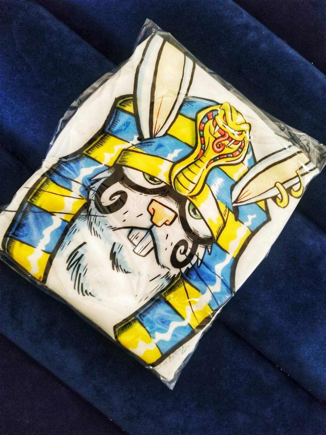 A colorful illustration of a rabbit with a unicorn horn, wearing a crown, on a pillow with a cat face design.