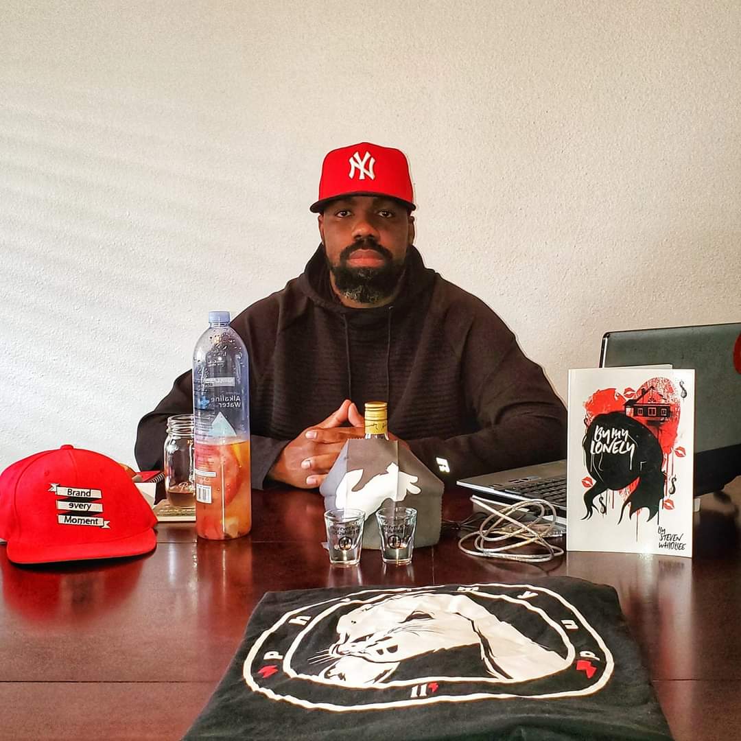 Man with beard wearing a red New York Yankees cap and black hoodie sitting at a table with various items, including a water bottle and a book.