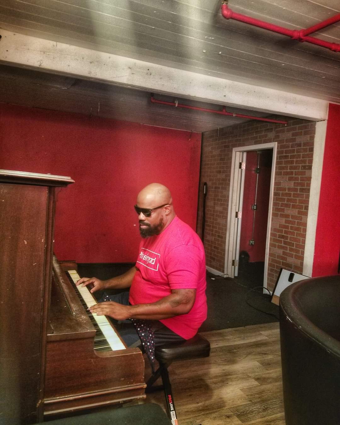 Person playing piano in a room with red walls and brick wall, wearing sunglasses and a pink shirt.