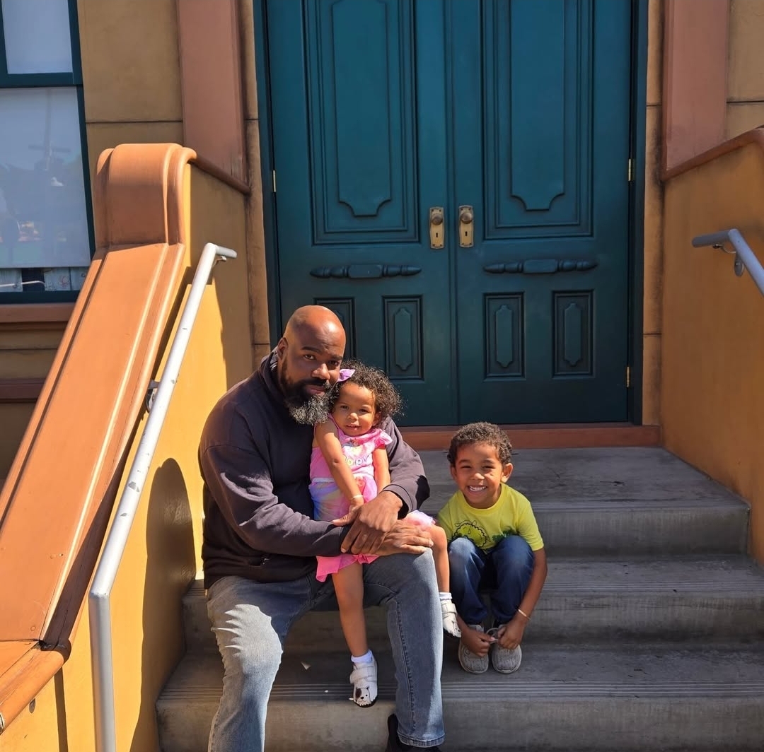 Man with two children sitting on stairs in front of a blue door, smiling, with colorful building walls around.