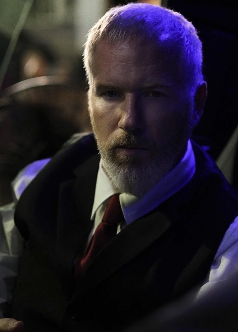Man with beard and short hair, wearing a suit and tie, in a dimly lit setting with blue and purple lighting.