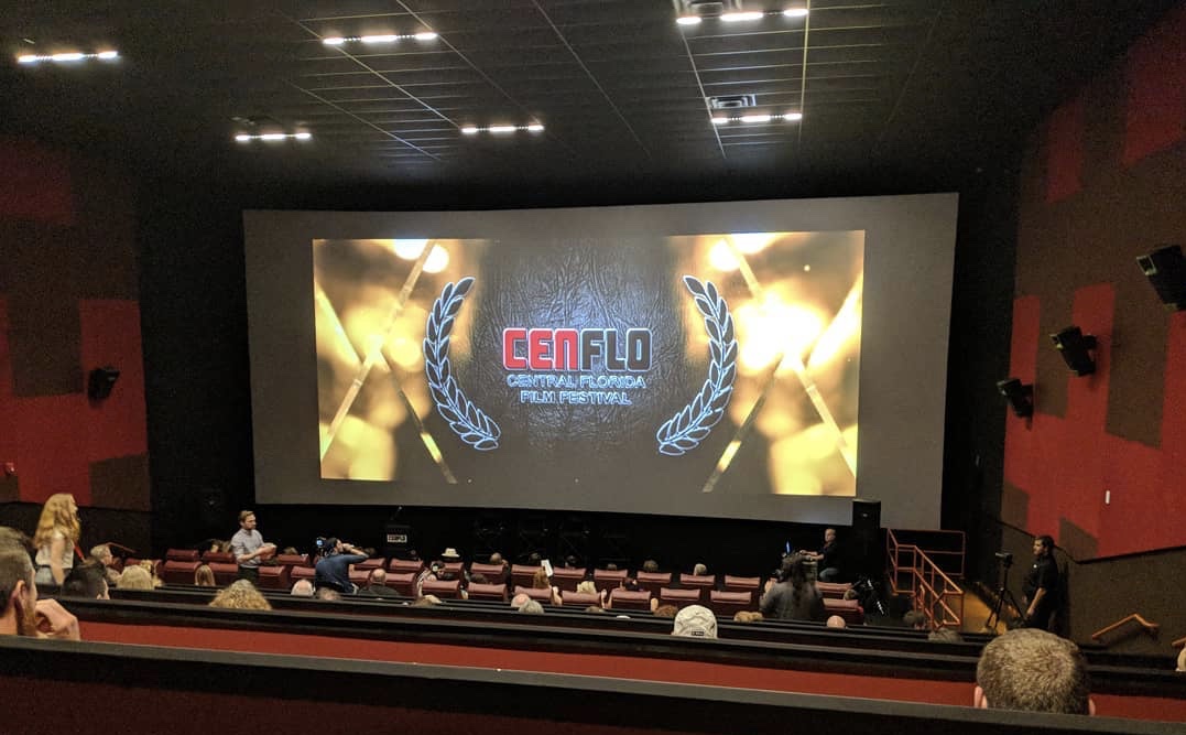 View of a movie theater with a large screen displaying a logo and people seated in the auditorium.