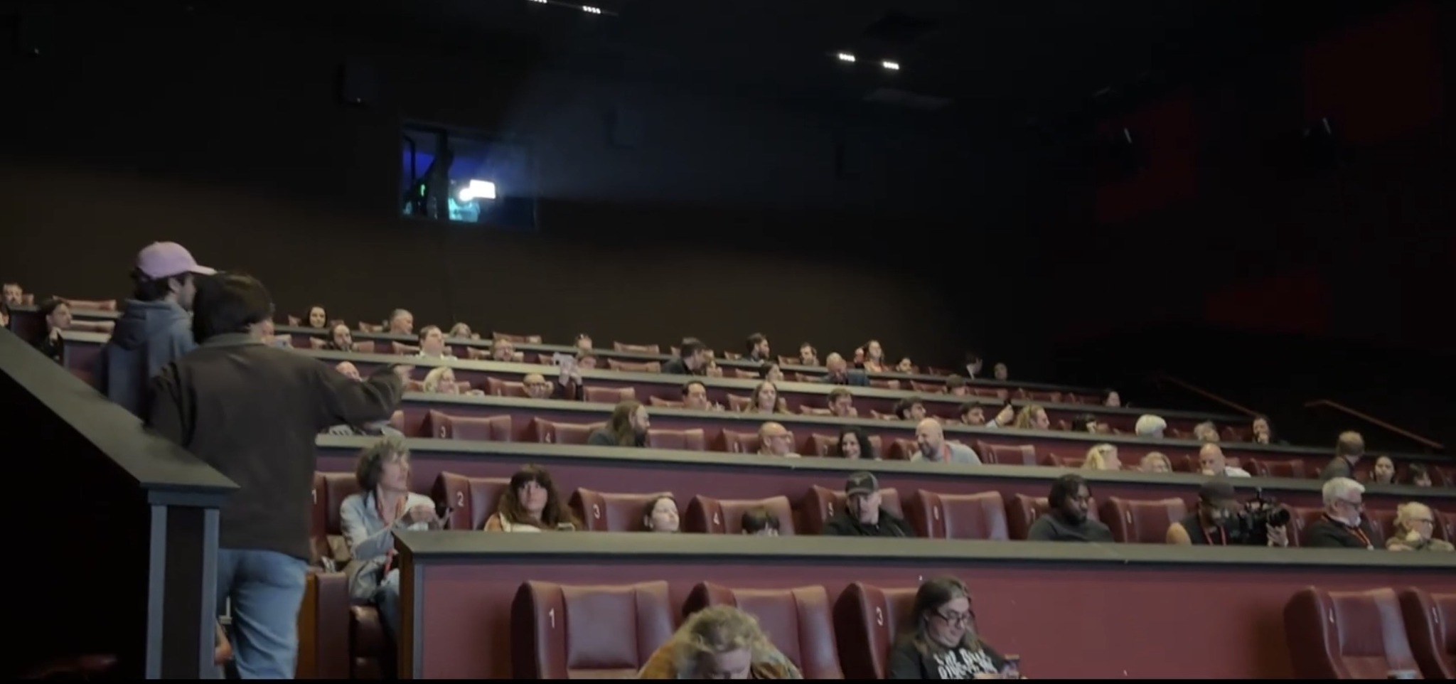 People seated in a large auditorium with multiple rows of seats, some standing and walking around, dim lighting.