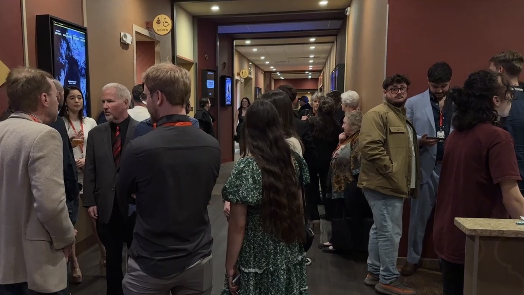People gathered in a hallway, engaging in conversations at an indoor event, with screens and decorations on the walls.