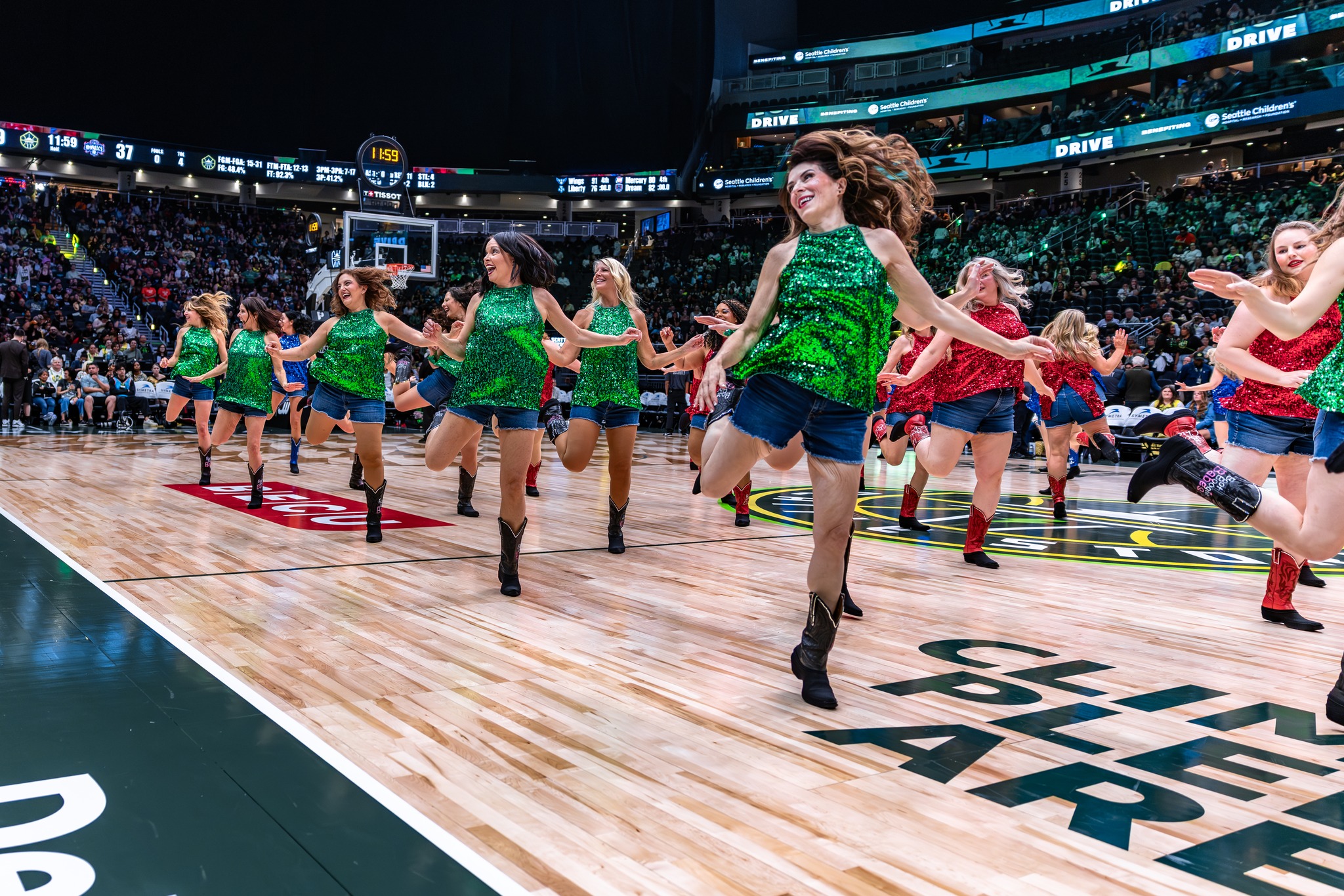 Group of dancers performing on a basketball court, wearing colorful costumes, with an audience in the background.