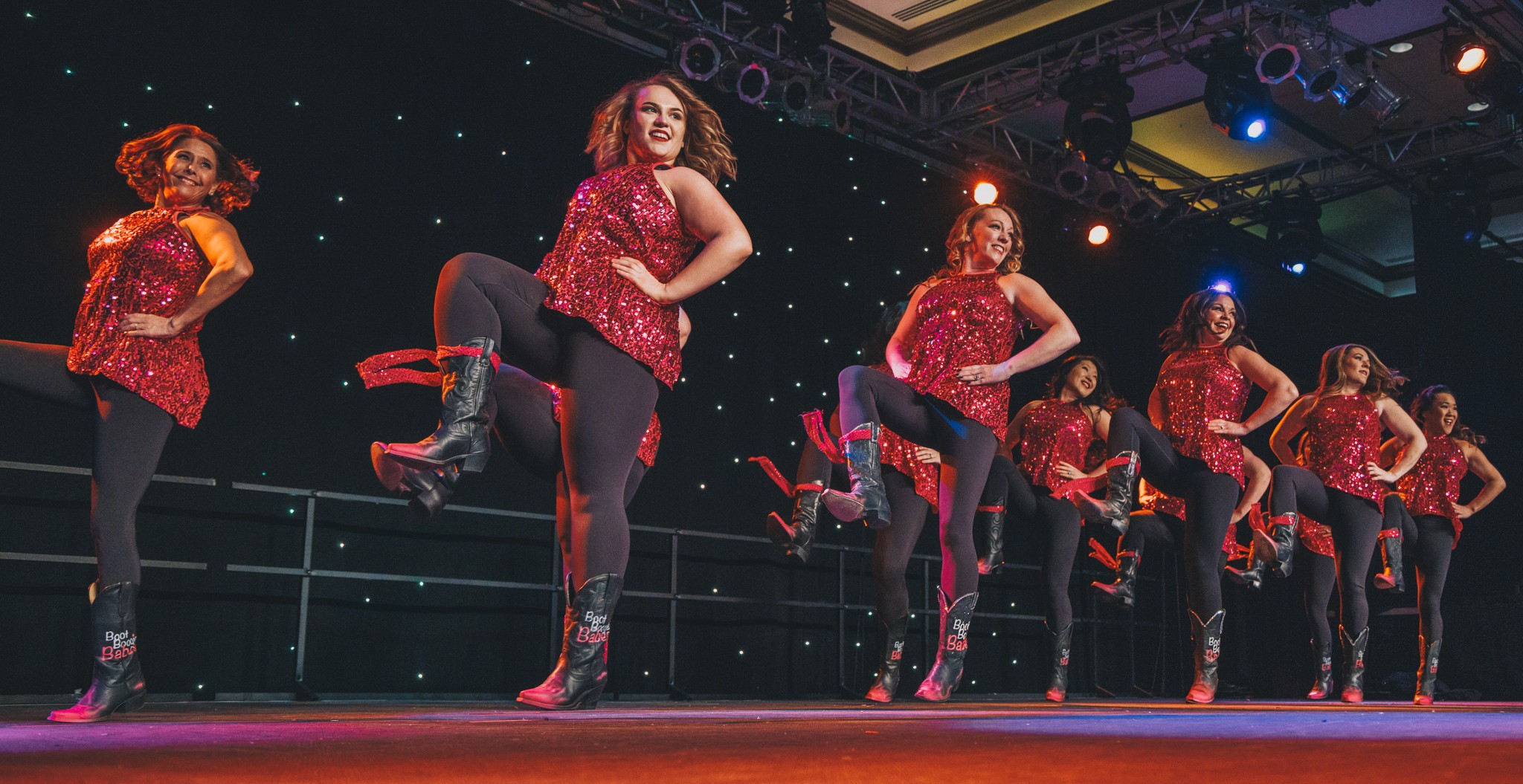 Group of dancers performing on stage in red costumes with black boots, mid-movement, stage lighting visible.