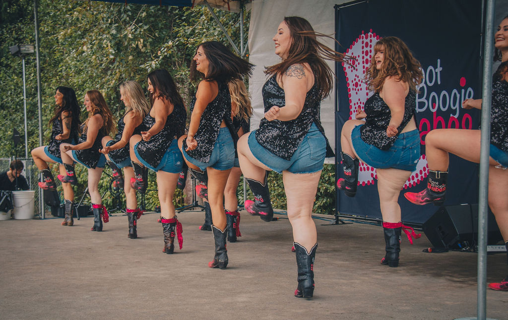 Group of women performing a dance outdoors, wearing matching outfits and boots, with trees and a banner in the background.