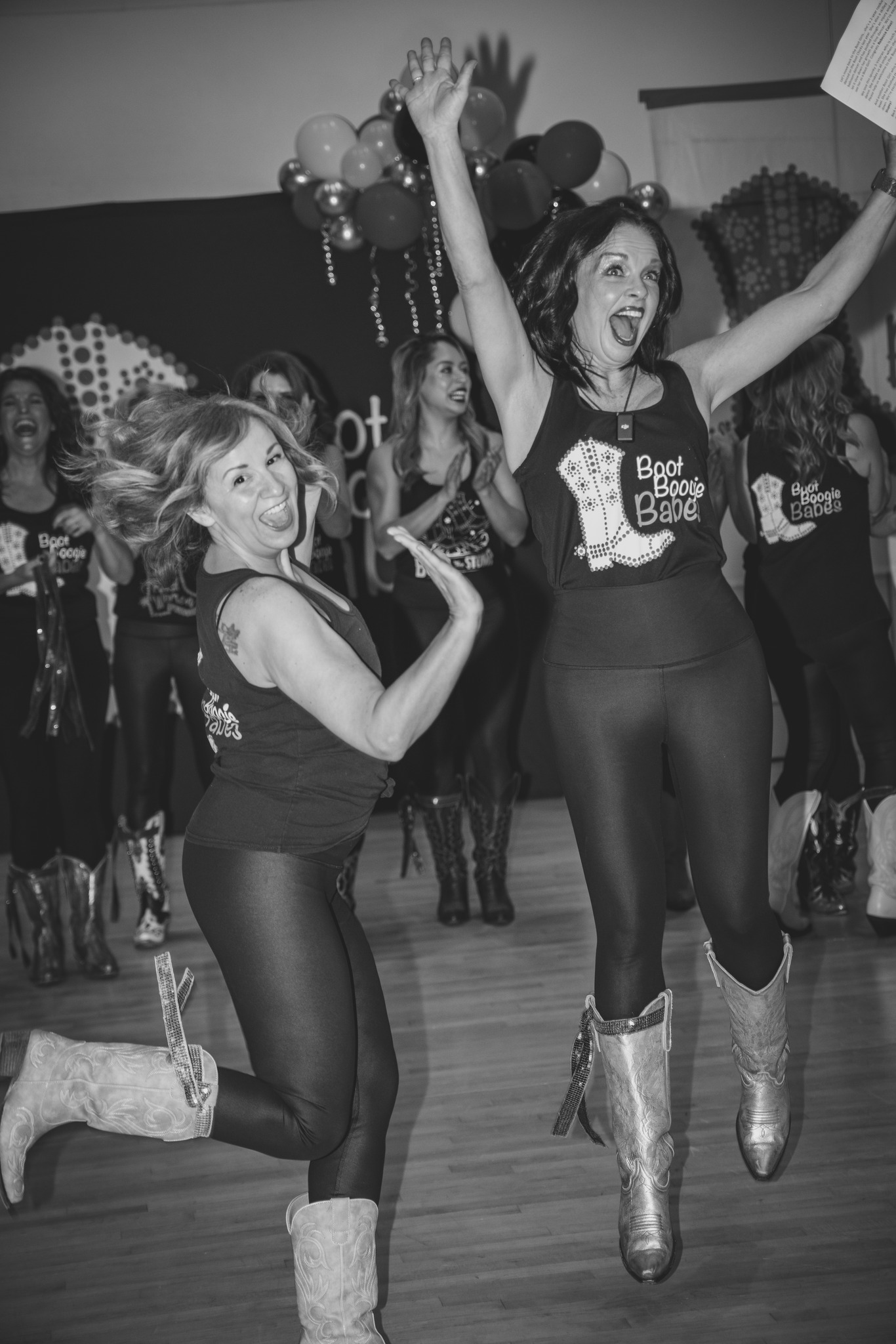 Two women dancing, one jumping with arms raised, others in background, balloons, and decorations, in a celebration setting.