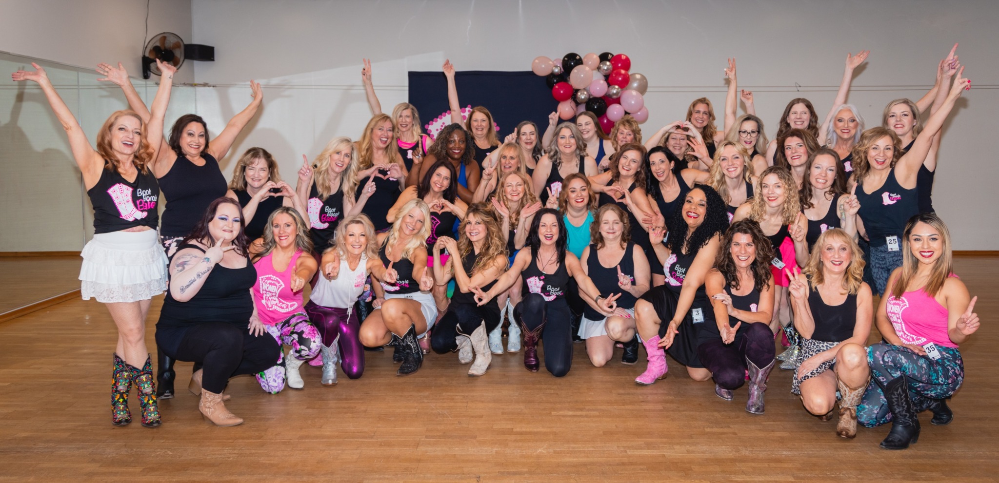 Group of women and girls posing together indoors, some with arms raised, in front of a decorated backdrop with flowers.