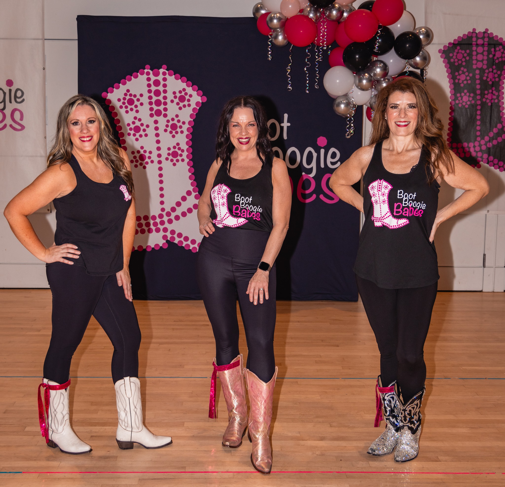 Three women in dance costumes stand on a stage with balloons and a backdrop, smiling at the camera.