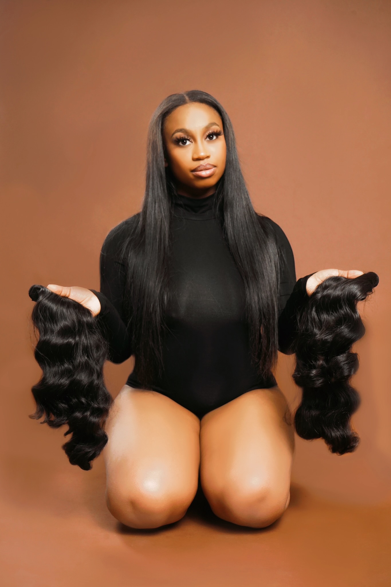 A woman with long black hair kneels on the floor, holding large sections of curly hair in each hand, against a brown background.