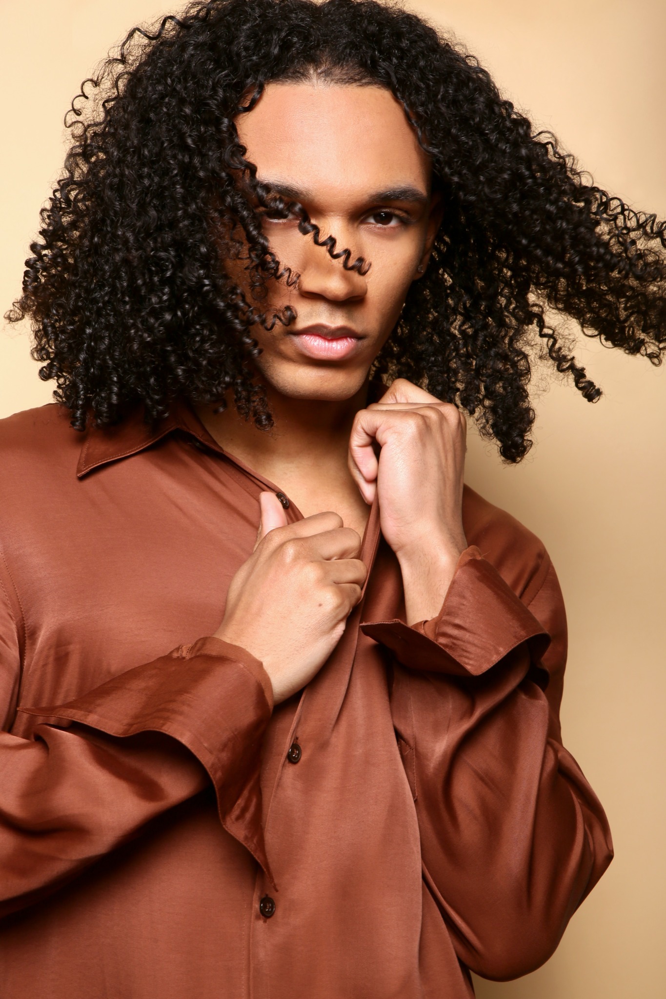 Woman with curly hair wearing a brown satin shirt, looking directly at the camera, hand near chin.