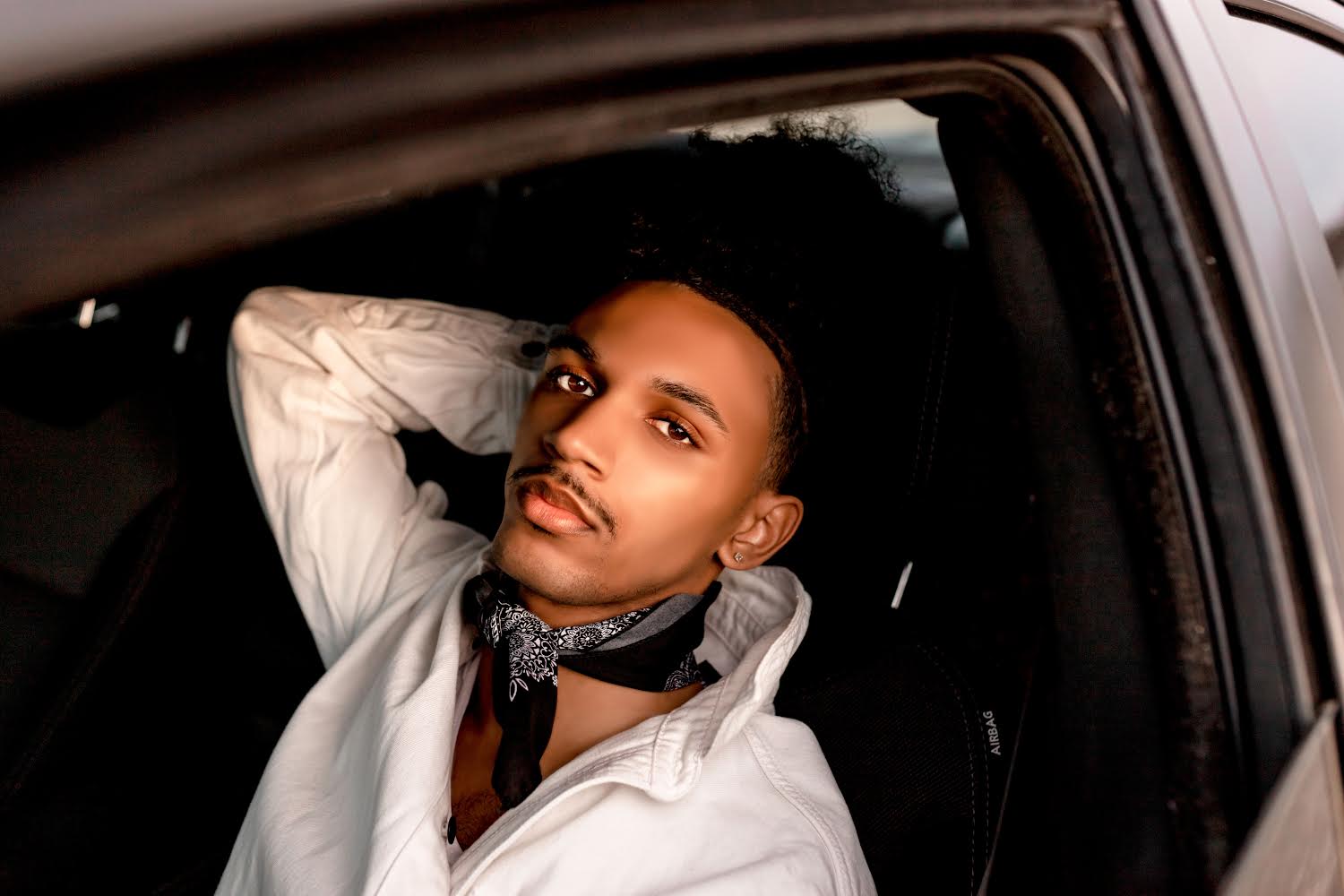 Young man with short hair and a mustache, wearing a white shirt and a patterned scarf, sitting in a car.