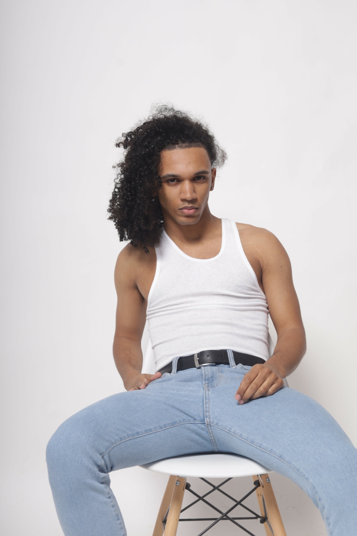 Person with curly hair wearing a white tank top and light blue jeans, sitting on a chair against a plain background.