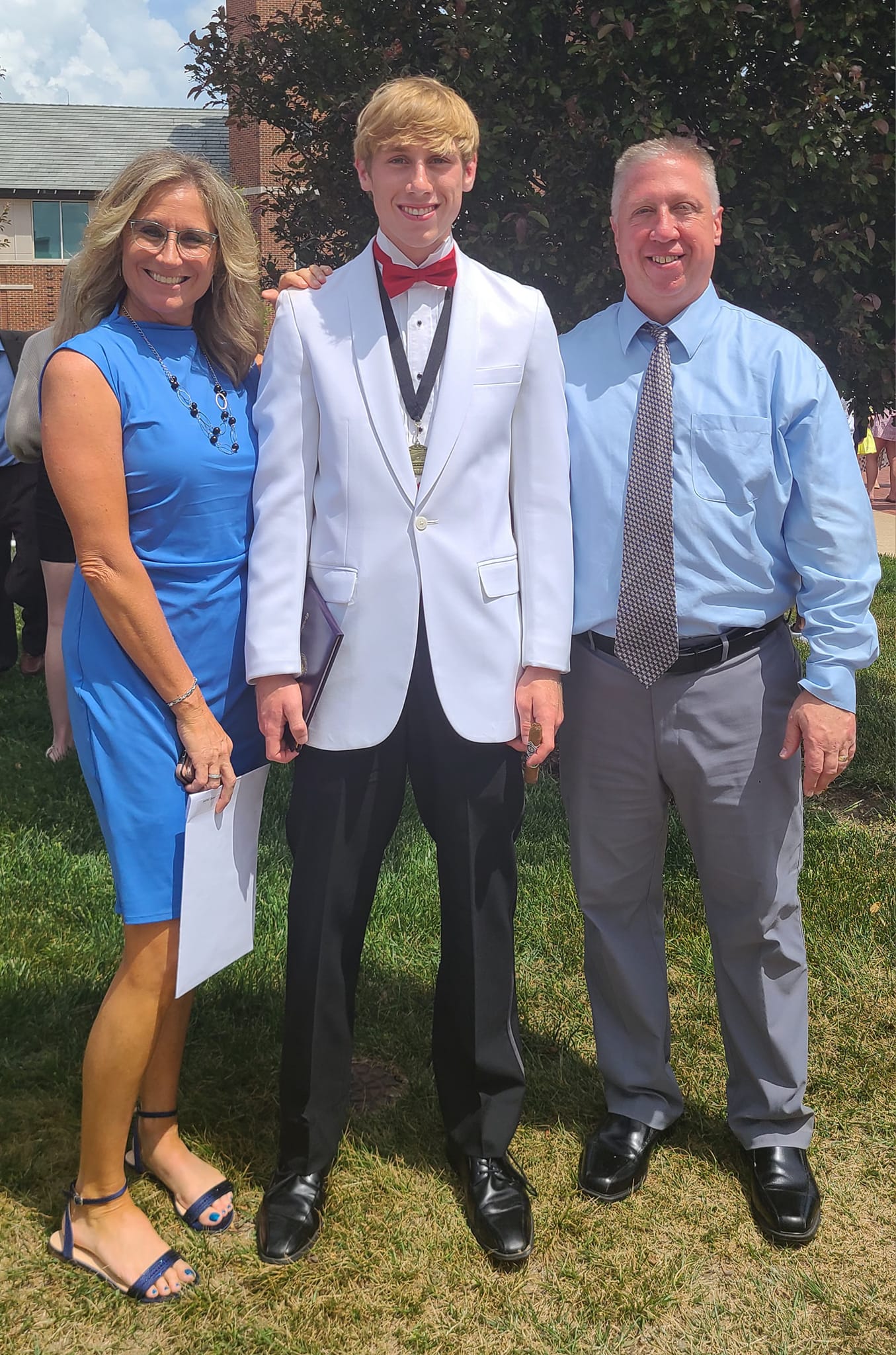 Three people standing outdoors, smiling, with a young man in a white suit and red bow tie in the center, flanked by a woman in a blue dress and a man in a light blue shirt and tie.