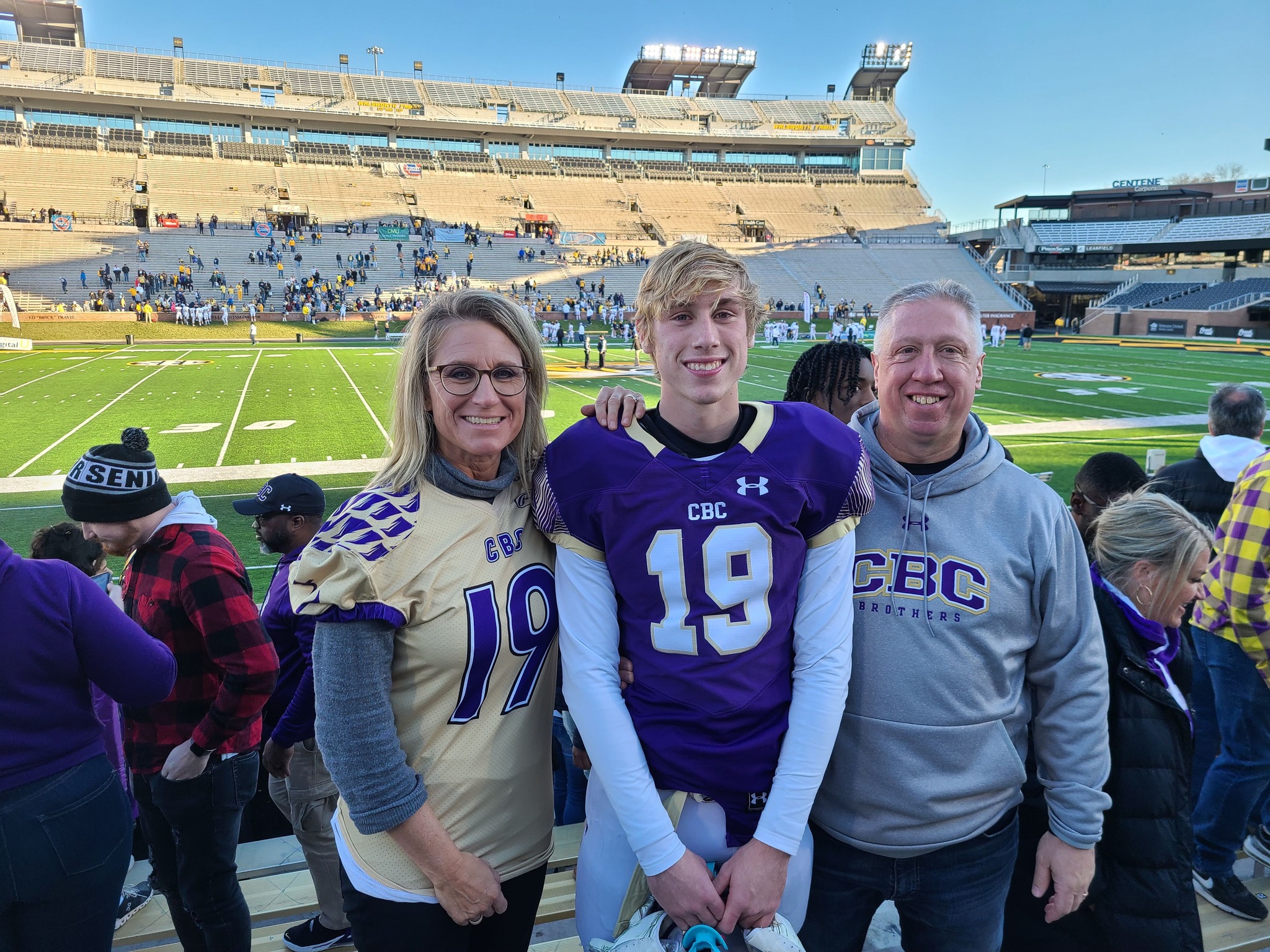 Three people standing on a football field with a stadium in the background, smiling, wearing purple and gray sports attire.