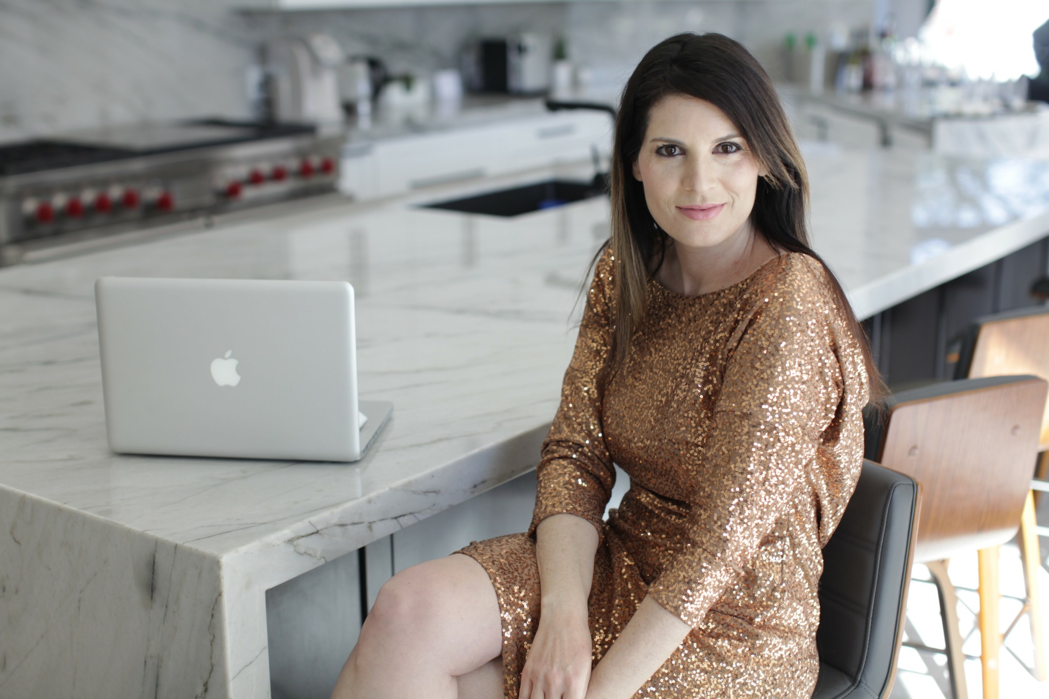 Woman sitting at a marble counter with a laptop in a modern kitchen, smiling, wearing a gold dress.