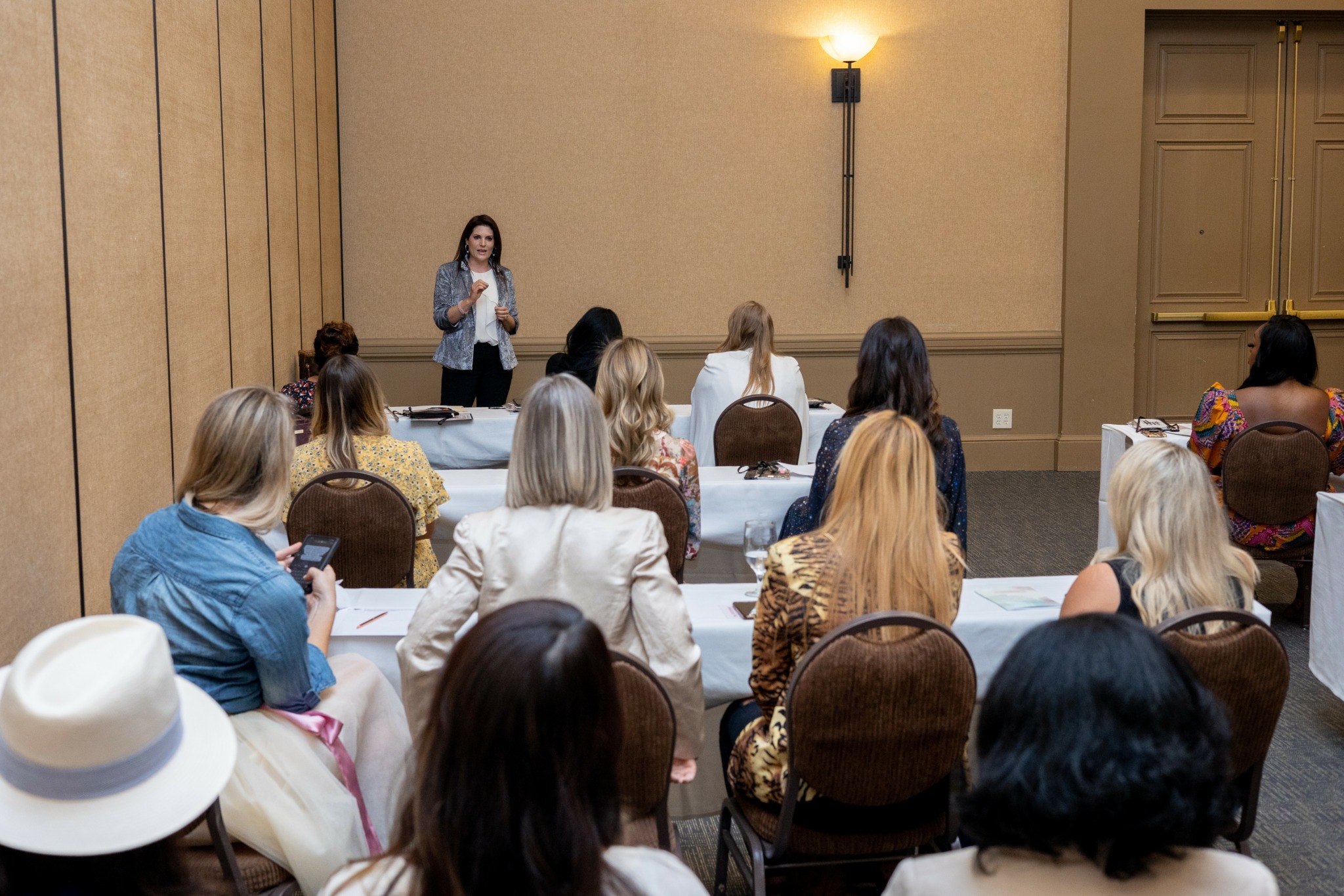 A woman stands at a podium speaking to an audience in a conference room with beige walls and a wall sconce.