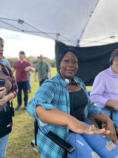 Group of people outdoors under a tent, woman in blue plaid shirt sitting, others standing nearby, grassy background