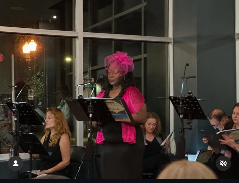 Woman with pink hair and pink top singing in a concert hall with musicians, large windows, and music stands.