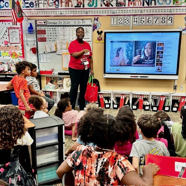 Teacher standing in front of a classroom with children seated, a large screen displaying a video call, colorful decorations, and educational materials.