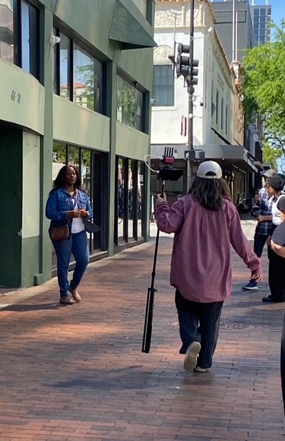 Person in pink jacket and white cap holding a camera on a tripod, walking on brick sidewalk, with people and buildings nearby.