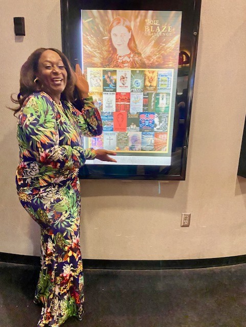 Woman in colorful floral outfit standing next to a digital display with book covers, smiling, in an indoor setting.
