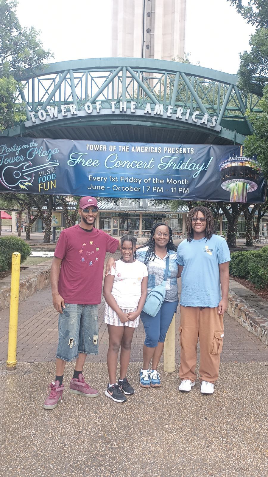 Four people standing in front of the Tower of the Americas entrance with a sign for a free concert event.