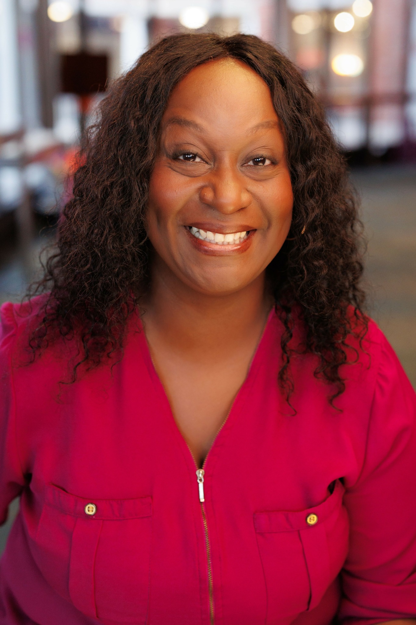 Smiling woman with curly hair wearing a red top, indoors with blurred background.