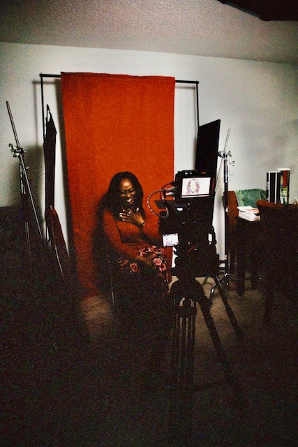 Person sitting in front of a camera setup with a red backdrop, in a dimly lit room.