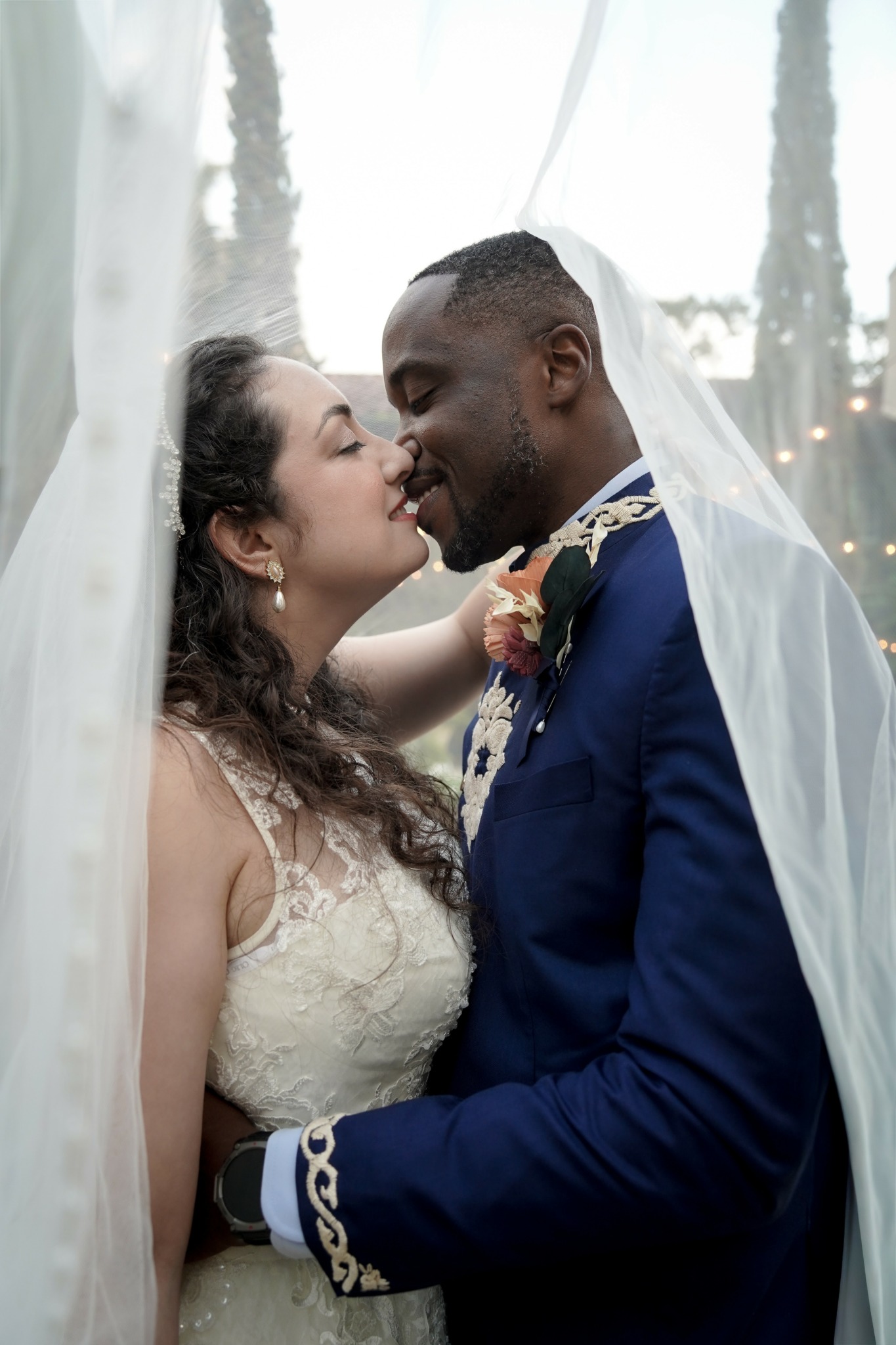 Bride and groom close together under a veil, smiling and touching noses, outdoors with trees in the background.