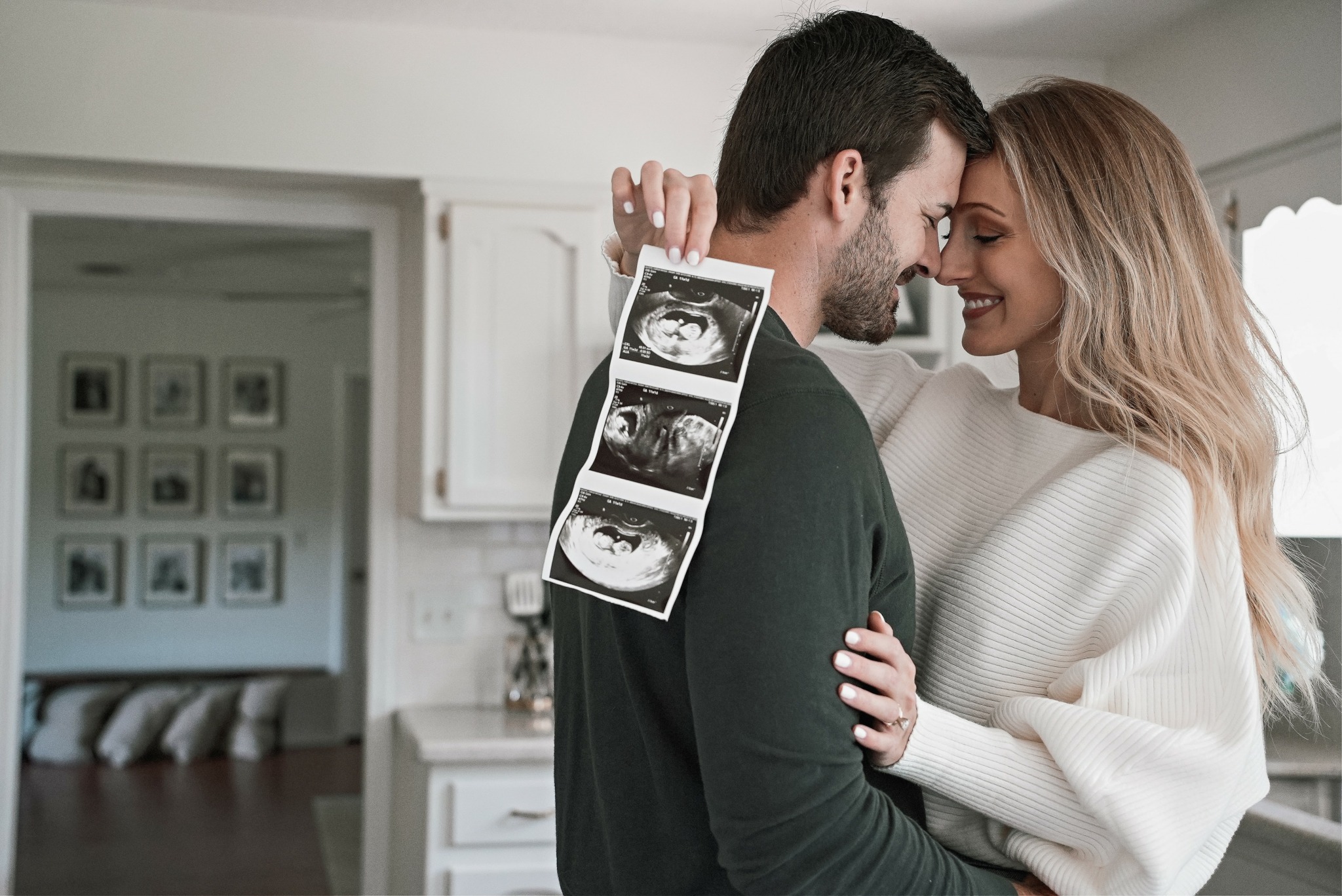 A couple smiling and touching foreheads, holding ultrasound images, in a bright kitchen. Woman has blonde hair, man has dark hair.