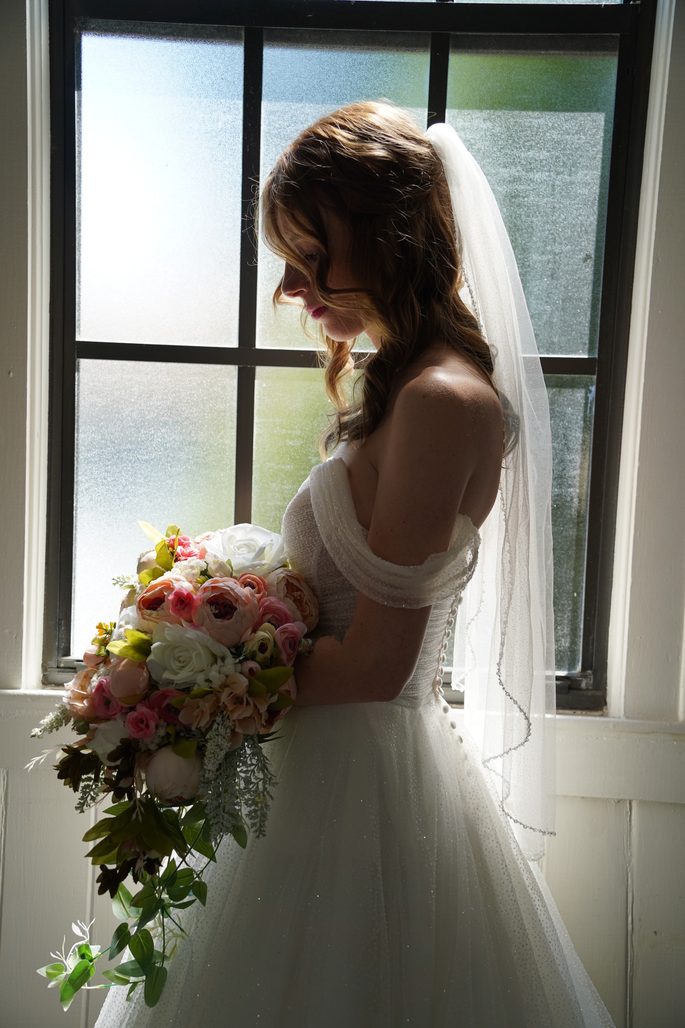 Bride holding a bouquet of flowers, standing by a window with sunlight, wearing a wedding dress and veil.