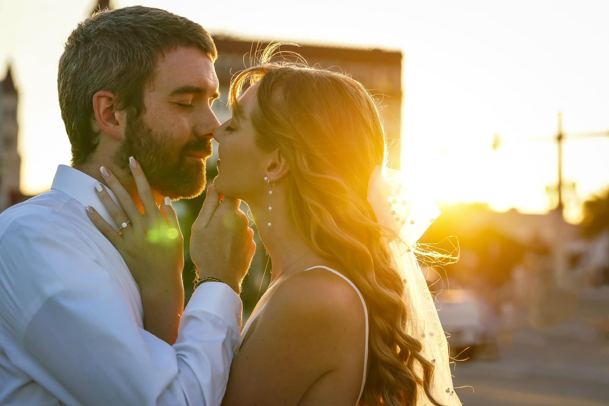 A man and woman face each other closely outdoors during sunset, with sunlight behind them, touching faces gently.