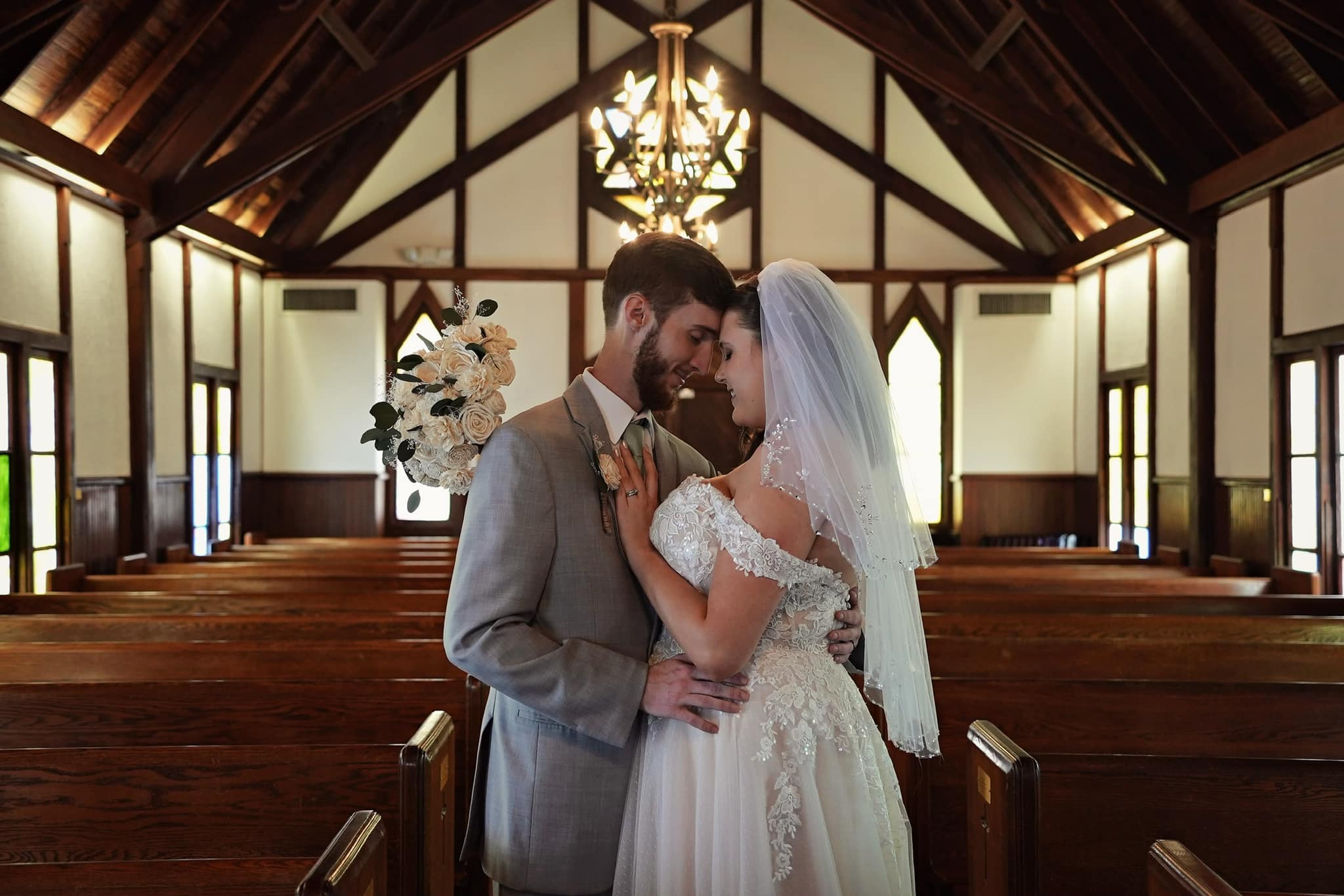 A bride and groom stand close together in a church, holding hands and smiling at each other, with a chandelier overhead.