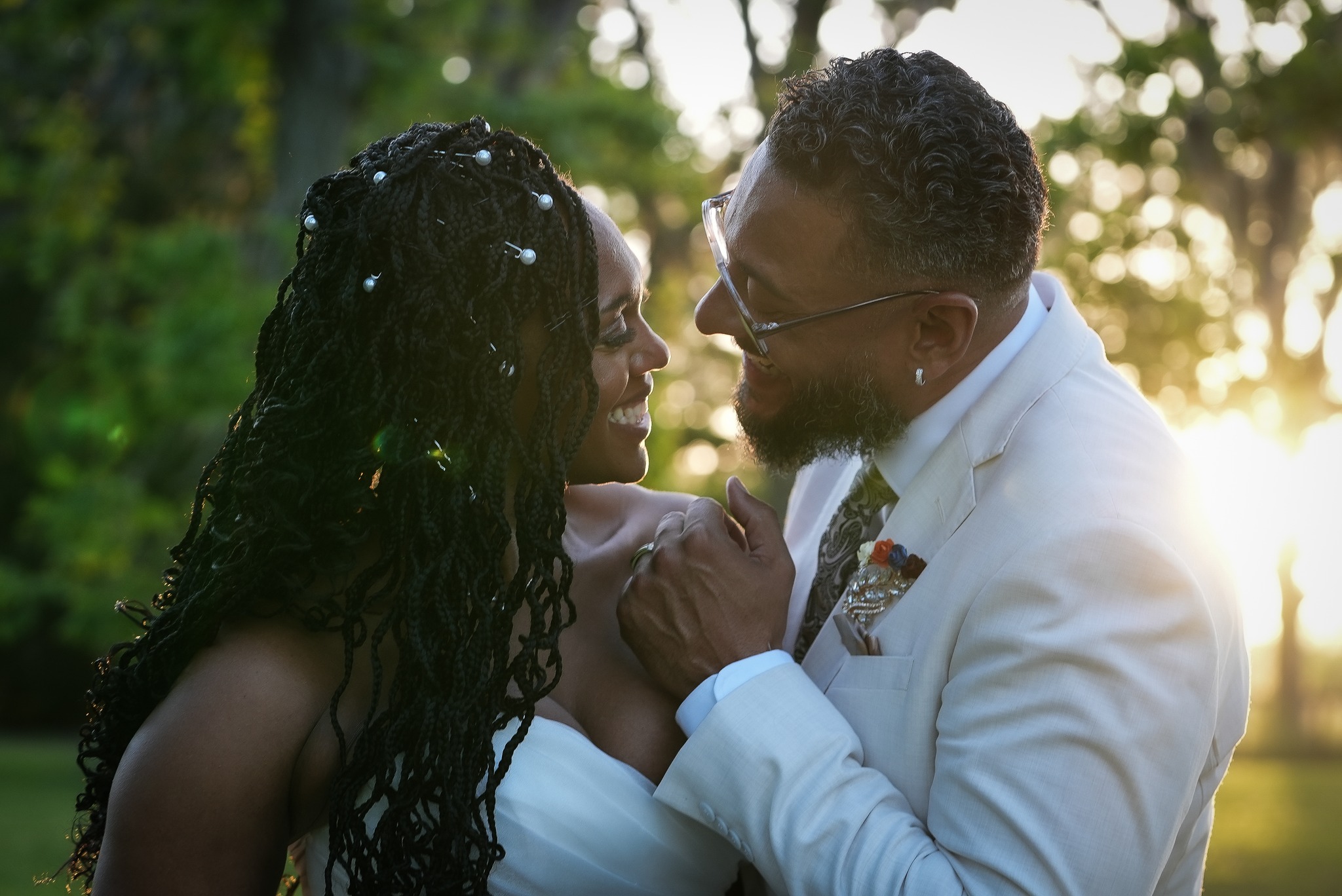 A couple faces each other outdoors with sunlight behind them, holding hands and smiling.